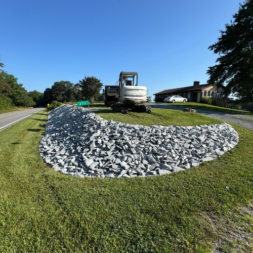 A small excavator sits on a lush green lawn beside a road, surrounded by a curved pile of gray stones, with a clear blue sky and trees in the background.