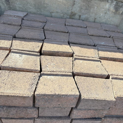 A stack of brown rectangular bricks neatly arranged in rows against a concrete wall. The bricks have a slightly uneven texture, suggesting an industrial setting.