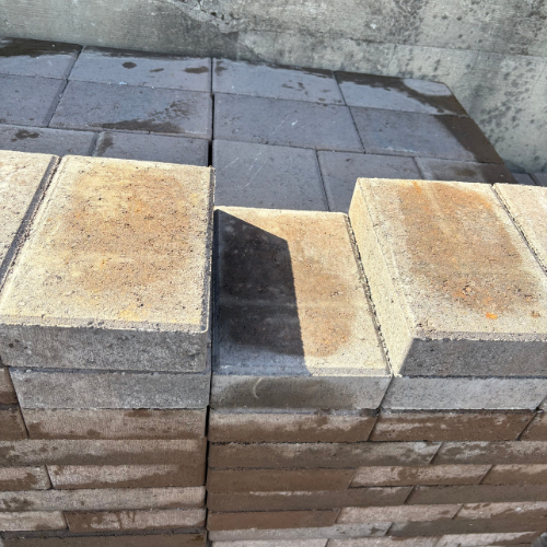 Stack of gray and beige concrete paving stones with rough, textured surfaces. They are neatly arranged against a concrete wall.