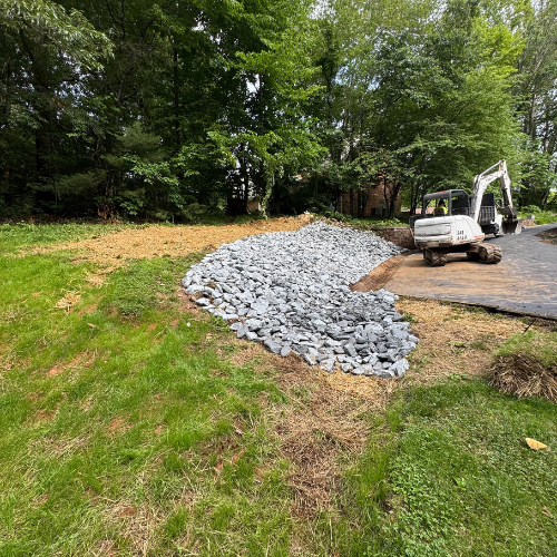 A small excavator near a newly laid gravel path curves through a lush, green lawn, bordered by dense trees, conveying a sense of progress and nature.