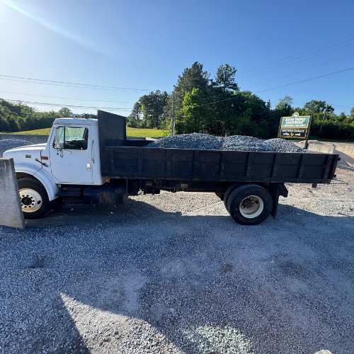 A white dump truck loaded with gravel is parked in a sunny lot surrounded by trees and a sign. The atmosphere is calm and industrious.