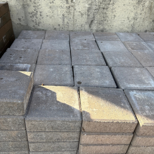 Stacked gray concrete pavers in sunlight, casting shadows. Positioned against a weathered concrete wall, creating a simple, industrial feel.