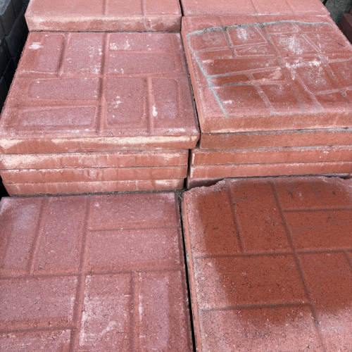 Stacks of red concrete paving slabs with geometric patterns. The slabs are slightly weathered, showcasing a mix of texture and uniformity.