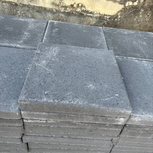 Stacked gray concrete pavers, showing smooth, square shapes with slight weathering. Background includes a weathered wall, conveying an industrial tone.