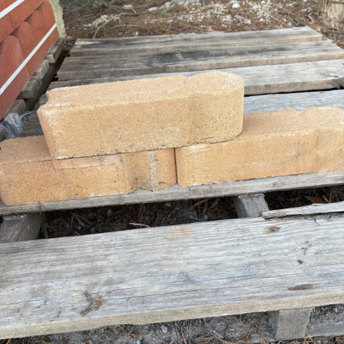 Three beige bricks are stacked on a wooden pallet outdoors, with red bricks visible to the side. The scene conveys simplicity and construction.