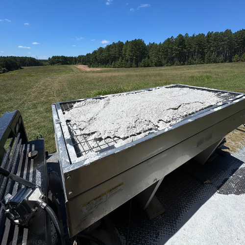 A metal container filled with a white, rocky substance sits outdoors on a sunny day. It is set in a grassy field with a backdrop of dense green trees.