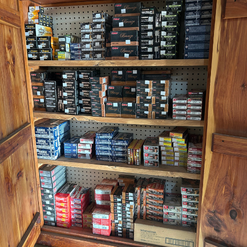 A wooden cabinet open to reveal shelves stacked with various boxes of ammunition, arranged neatly by type and size, against a pegboard backdrop.