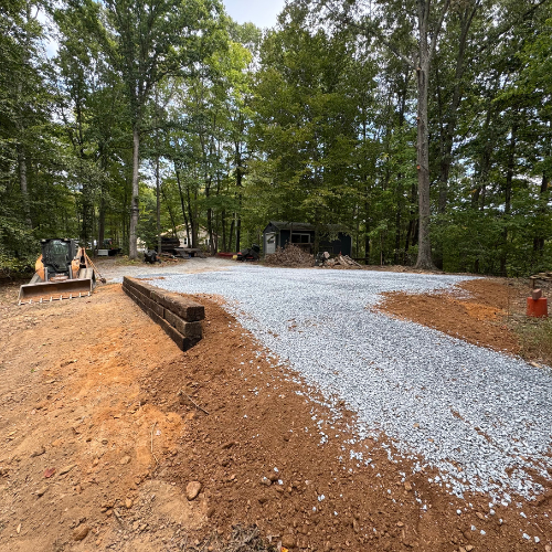 A gravel driveway curves through a wooded area with tall trees and a small shed. A bulldozer and timber line the path, suggesting ongoing construction.