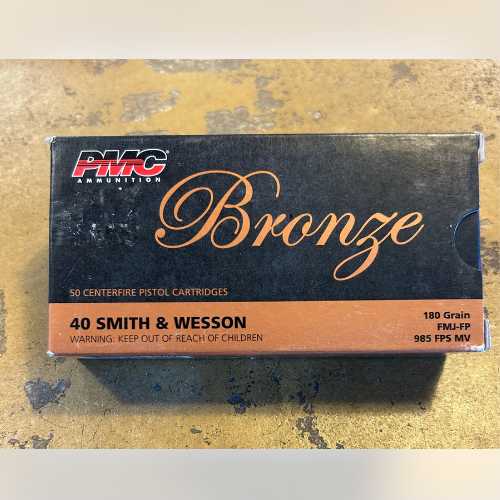 A box of PMC Bronze ammunition on a textured surface. The box labels it as .40 Smith & Wesson, 180 Grain FMJ-FP, with a child safety warning.