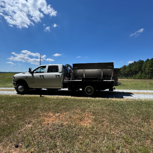 A white utility truck with equipment on the back is parked on a gravel path, surrounded by a grassy field under a clear blue sky with scattered clouds.