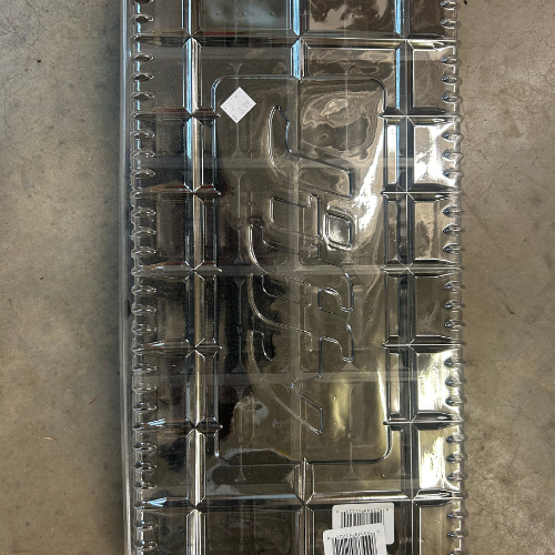 Transparent plastic tray with a grid design on a gray surface, featuring embossed text and a barcode sticker, reflecting light subtly.