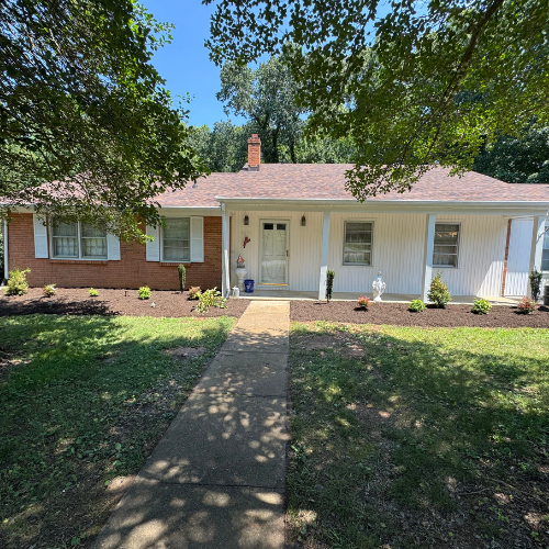 A charming one-story brick house with red roof, white siding, and a central chimney. Neat landscaping and a shaded lawn create a peaceful atmosphere.