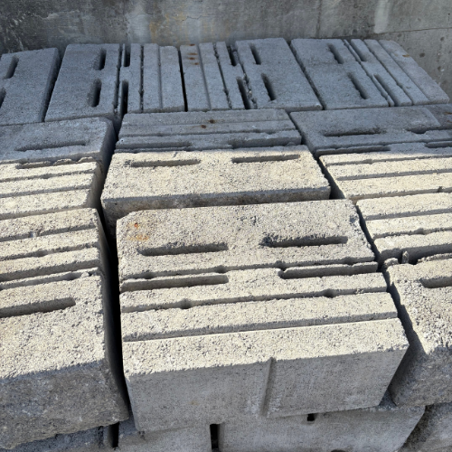 A stack of gray concrete blocks with grooves, arranged neatly in a pile. The surface texture appears rough, set against a concrete wall background.
