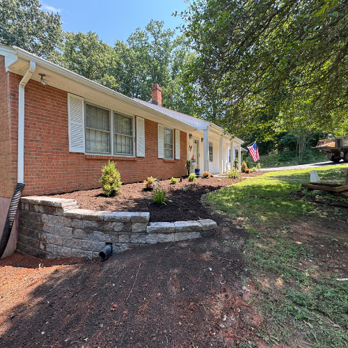 Single-story brick house with white shutters, surrounded by a neatly landscaped garden and American flag. Bright, sunny day enhances greenery.