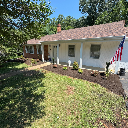 Single-story house with a red brick and white facade, surrounded by manicured landscaping. An American flag is displayed near the entrance. Calm, sunny day.