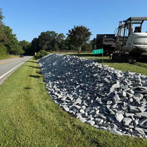A large pile of gray rocks lines a road with green grass on the side. Construction vehicles are parked nearby. A green sign displays the number 373.