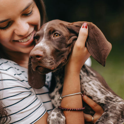 Pet Food & SuppliesWoman smiling while holding a brown puppy with floppy ears. She wears striped shirt and bracelets. The scene conveys warmth and affection outdoors.