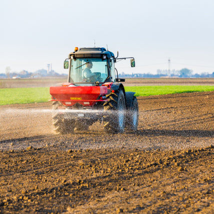 FertilizersA red tractor sprays fertilizer over a brown plowed field under a clear sky, with green crops in the background, symbolizing farming productivity.